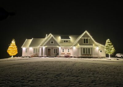 A house with a snow-covered lawn is adorned with christmas lights along the roof and two lit trees in yellow and white, showcasing a beautiful holiday light installation at night.