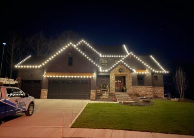 A house at night with holiday lights outlining the roof and garage; a work truck is parked in the driveway, suggesting a recent holiday light installation.