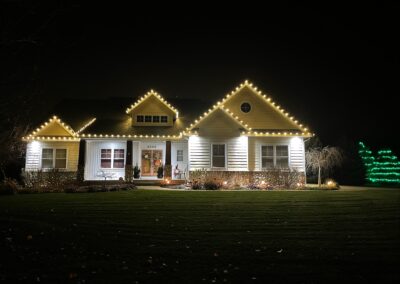 A house at night with white and yellow holiday lights outlining the roof and porch. A tree on the right side is decorated with green lights, showcasing a festive holiday light installation.