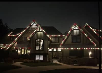 A large house at night with red, green, and white christmas lights outlining the roof and gables, creating a festive holiday lights display.