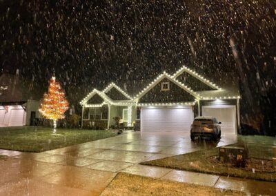 Single-story house with professional holiday light installation outlining the roof, a decorated tree in the yard, parked car in the driveway, and snow falling at night.