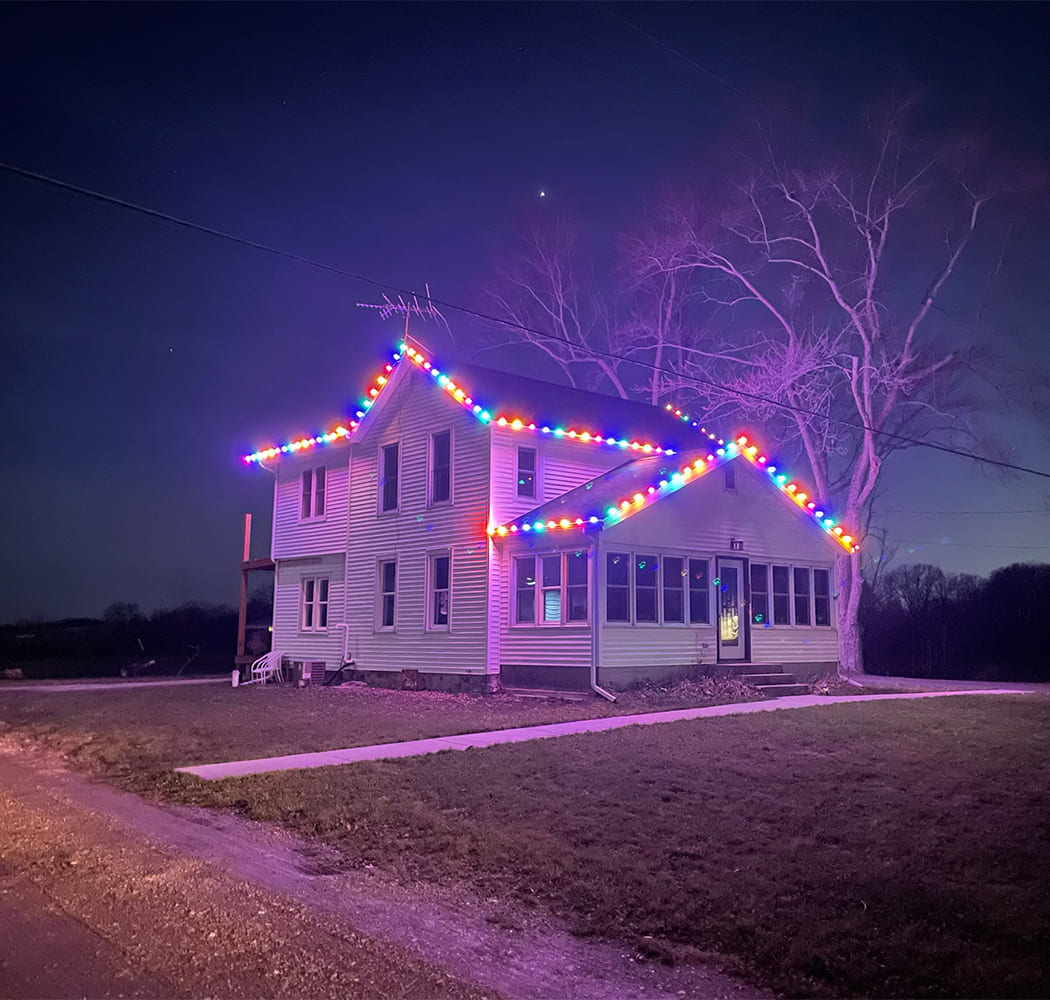 A two-story house decorated with colorful holiday lights along the roofline at night, with bare trees and a dark sky in the background.