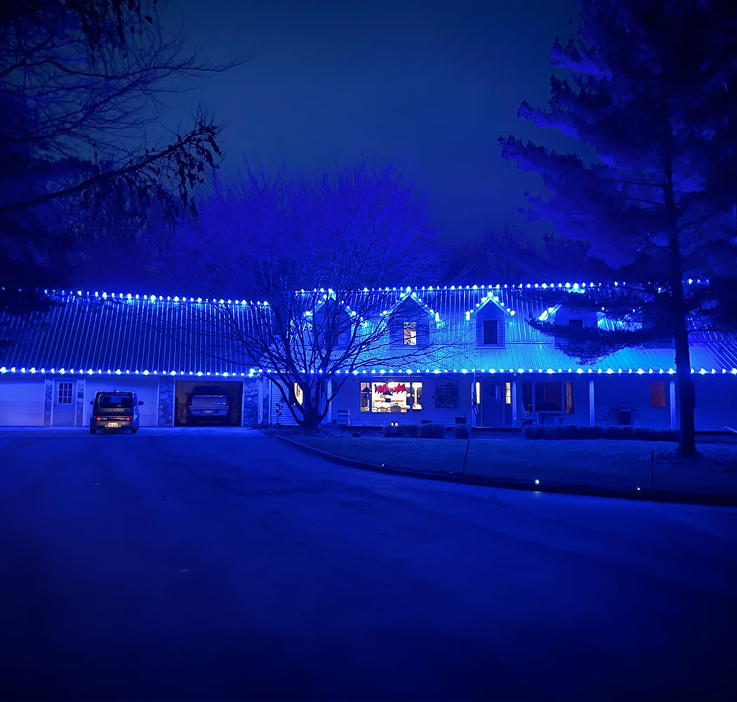 A house with a wraparound porch features bright blue holiday lights along the roofline at night, showcasing a stunning holiday light installation; a car is parked on the left in the driveway.