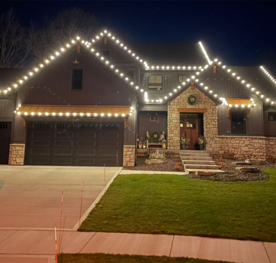 A two-story house at night with stone and wood exterior, decorated with white holiday lights along the roofline, and a wreath on the front door.