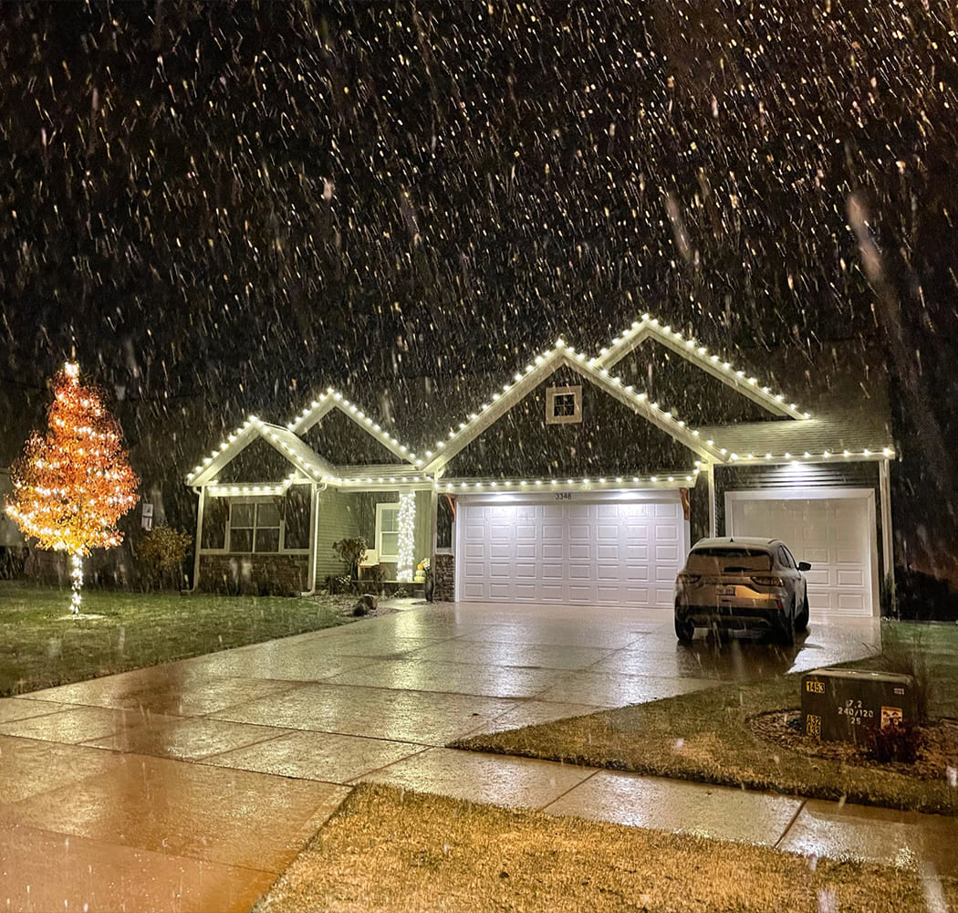 Single-story house with glowing holiday lights and a lit tree in the yard at night, as snow falls gently and a car is parked in the driveway.