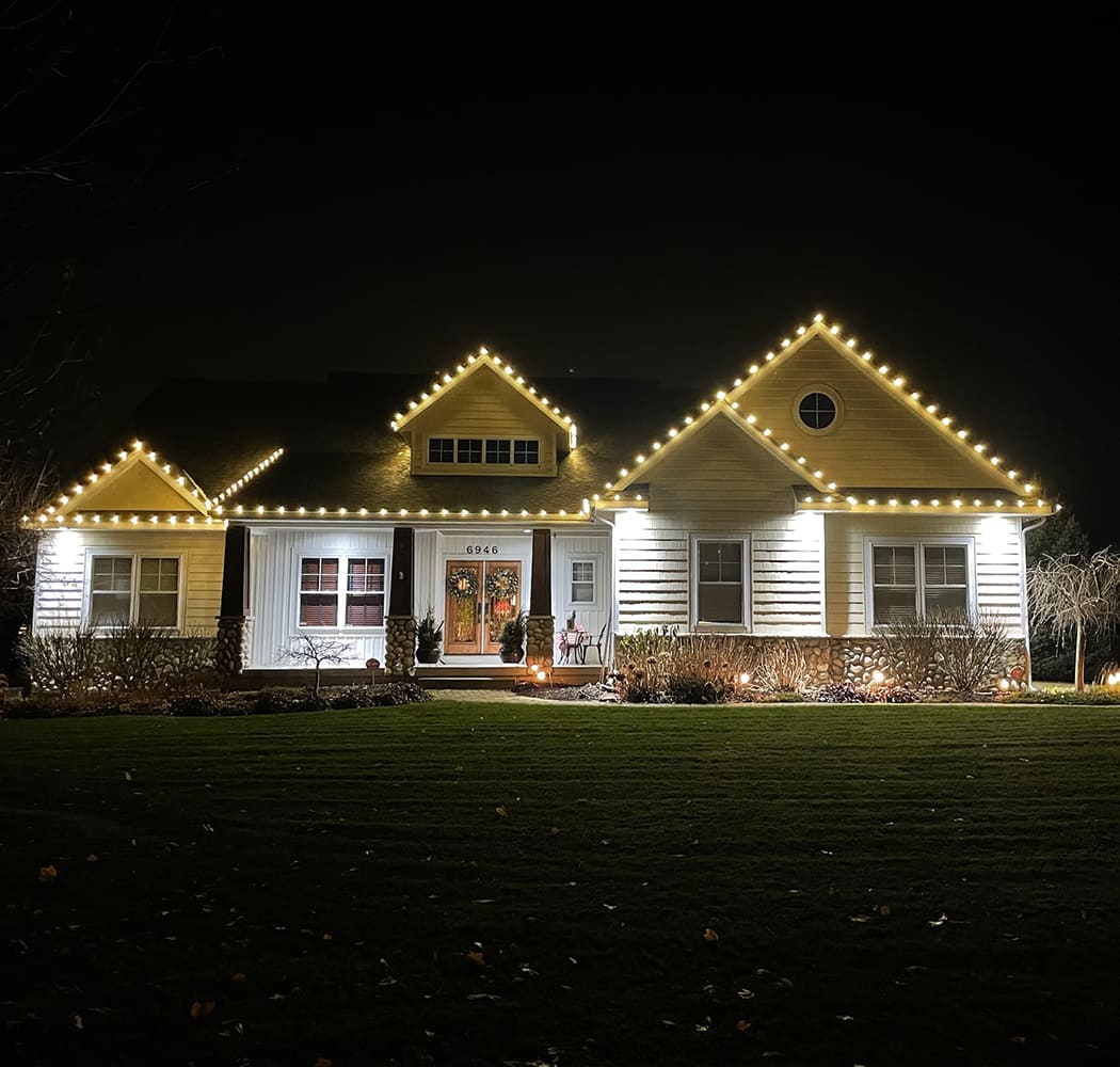 A single-story house features a beautiful holiday light installation, with white string lights outlining the roofline and glowing at night, while a lawn is visible in the foreground.