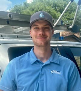 A man wearing a blue polo shirt and cap with a "Pure Pro Cleaning" logo stands in front of a white vehicle with a ladder on top, ready for christmas light installation, outdoors on a sunny day.